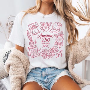 May include: White t-shirt with a red graphic design celebrating America's 250th anniversary. The design includes an eagle, flowers, a bell, a quill, and the words "America 250" with the years 1776-2026.