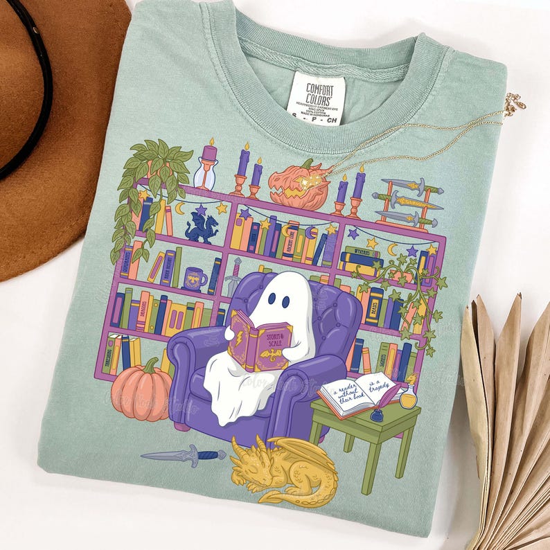 May include: A light green Comfort Colors t-shirt with a fantasy-themed design. A ghost reads a book in a purple armchair in front of a bookshelf. The bookshelf is filled with books, candles, and a pumpkin. A golden dragon and a small table with a quill and ink are also present.