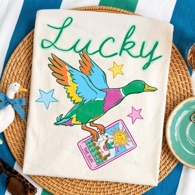 May include: Cream-colored t-shirt with a colorful duck graphic, the word "Lucky" in green script, and star accents. The duck holds a tarot card that reads "The Sun". The shirt is on a woven mat.