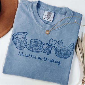 Peut inclure: T-shirt bleu clair Comfort Colors avec un motif graphique représentant une cruche, une tasse à thé, des cuillères et une poule dans un panier. Le texte "I'd rather be thrifting" est imprimé en dessous. Un collier doré est posé sur le t-shirt.