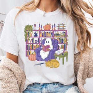 May include: White t-shirt with a colourful illustration of a ghost reading in a purple armchair. The design includes a bookshelf, a pumpkin, a sleeping dragon, and a small table with a book and quill.
