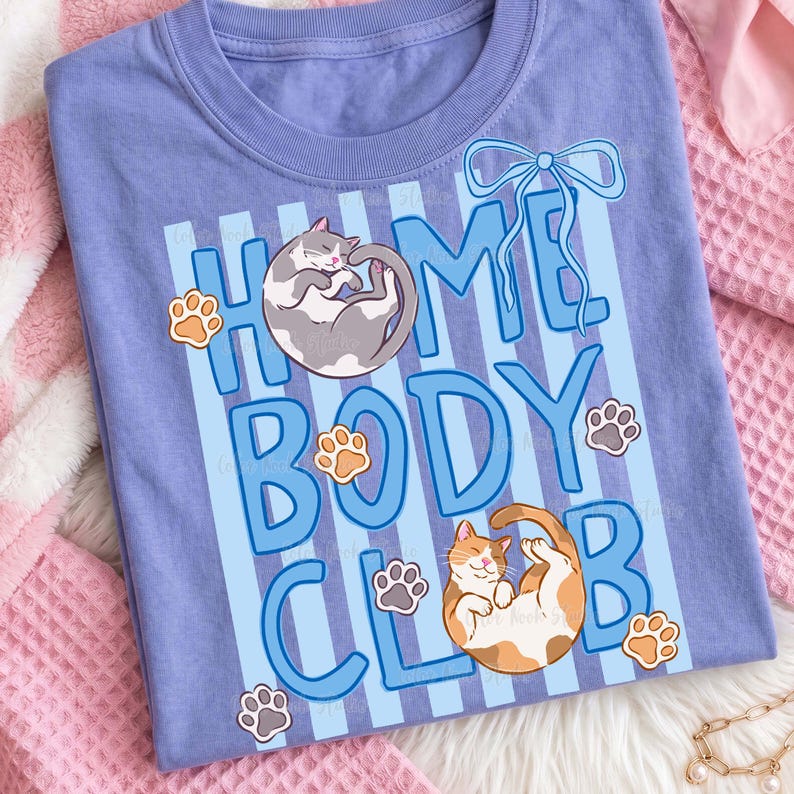 May include: A light blue t-shirt with the words "HOME BODY CLUB" in blue block letters. The shirt features illustrations of two cats and paw prints. The cats are gray and orange. The shirt is made of soft material.