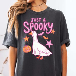 Może przedstawiać: Ciemnoszara koszulka z napisem "Just a Spooky" w kolorze r&oacute;żowym. Przedstawiono białego, upiornego gęsia z pomarańczowymi stopami i dziobem, a także pomarańczowe gwiazdy, nietoperze i dynię. Koszulka ma kr&oacute;tkie rękawy.