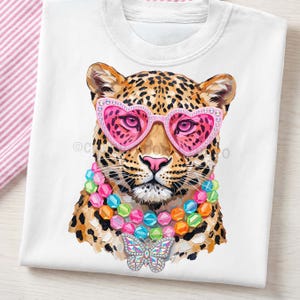 May include: A white t-shirt featuring a graphic illustration of a leopard wearing pink heart-shaped sunglasses with rhinestones, a multi-colored beaded necklace, and a large, iridescent butterfly pendant hanging from the center of the necklace.