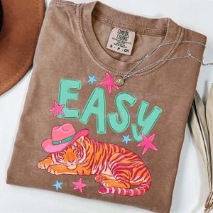 Puede incluir: Camiseta Comfort Colors marr&oacute;n con un gr&aacute;fico de un tigre con un sombrero de vaquero rosa, la palabra "EASY" en turquesa y estrellas rosas y azules. Un collar plateado con un colgante est&aacute; sobre la camiseta.