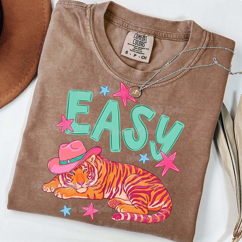 Puede incluir: Camiseta Comfort Colors marr&oacute;n con un gr&aacute;fico de un tigre con un sombrero de vaquero rosa, la palabra "EASY" en turquesa y estrellas rosas y azules. Un collar plateado con un colgante est&aacute; sobre la camiseta.