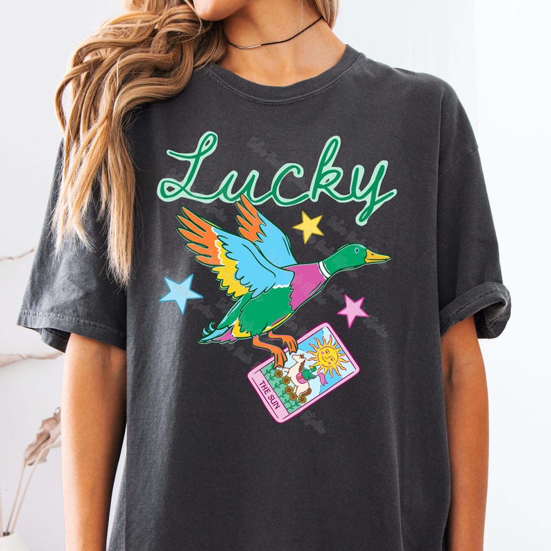 May include: Dark gray t-shirt featuring a colorful duck graphic with the word "Lucky" in green script. The duck has blue, orange, green, and pink feathers and is holding a tarot card. Stars in blue, yellow, and pink surround the duck.