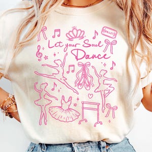 May include: Cream-colored sweatshirt with a pink ballet-themed graphic. The design includes ballerinas, ballet shoes, musical notes, a crown, and the text "Let your Soul Dance". A ballet ticket and bows complete the design.