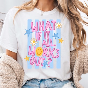 May include: White t-shirt with the text "WHAT IF IT ALL WORKS OUT?" in pink, with blue stars, yellow stars, and daisy accents. The shirt has blue vertical stripes in the background.