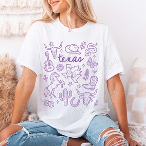 May include: White t-shirt with a purple Texas-themed graphic. The design includes a guitar, cowboy hat, longhorn skull, horse, cactus, and the word "Texas". The shirt is a relaxed fit.