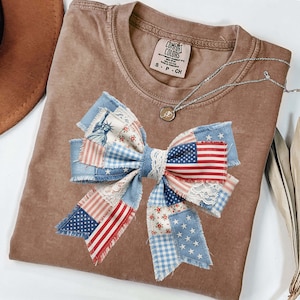 May include: A brown Comfort Colors t-shirt featuring a large patchwork bow design. The bow incorporates denim, floral, and American flag patterns, with a lace center. A silver necklace with a small pendant is also visible.