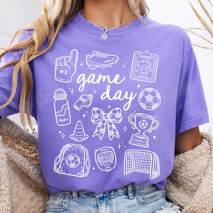 May include: Purple t-shirt with a white graphic design that says "game day" and includes various sports-related icons, including a football, a whistle, a water bottle, a trophy, and a pair of boots.
