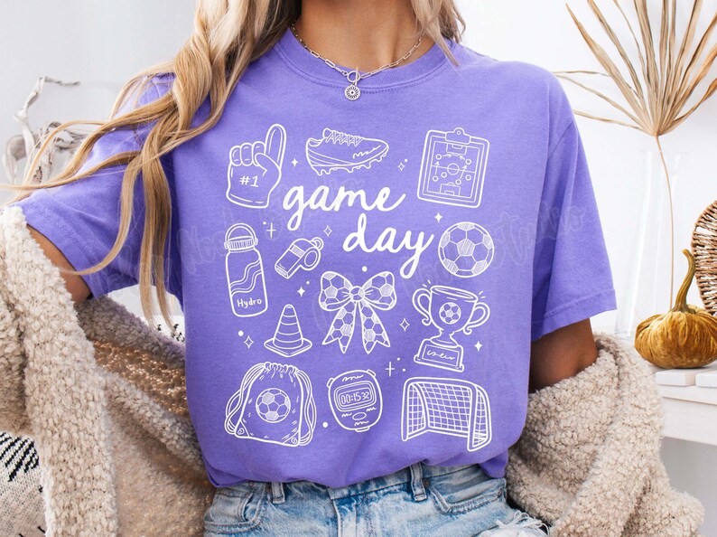 May include: Purple t-shirt with a white graphic design that says "game day" and includes various sports-related icons, including a football, a whistle, a water bottle, a trophy, and a pair of boots.