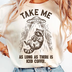 May include: Cream-colored t-shirt with a graphic of a raccoon being beamed up by a UFO, holding an iced coffee. Text above reads "TAKE ME" and below "AS LONG AS THERE IS ICED COFFEE."