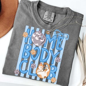 May include: A gray Comfort Colors t-shirt with the words "HOME BODY CLUB" in blue, featuring illustrations of two cats and paw prints. A silver necklace with a pendant is on the shirt.