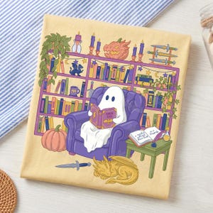May include: A beige cushion featuring a cartoon ghost reading a book in a purple armchair in front of a bookshelf. The design includes a pumpkin, a sleeping dragon, and the text "STORIES & SCALES".