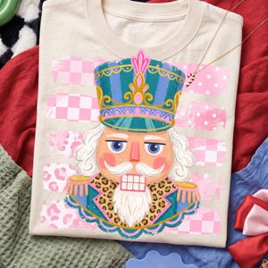 May include: A cream-coloured t-shirt featuring a graphic of a nutcracker wearing a teal and gold uniform with a leopard print collar. The background design includes horizontal stripes of pink and white checkered and polka dot patterns.