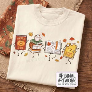 Può includere: Una t-shirt color crema con una grafica di quattro libri antropomorfi con facce sorridenti. I libri sono etichettati Pumpkin Spice, Cozy Reads e Autumn Classics, accompagnati da una tazza di cacao, foglie autunnali e un paio di occhiali.