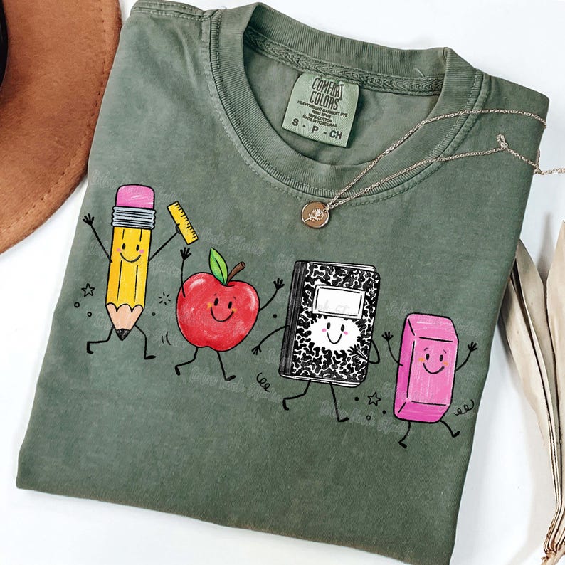 May include: Sage green Comfort Colors t-shirt featuring a playful graphic of animated school supplies: a pencil, apple, notebook, and eraser. The shirt has a crew neck and short sleeves. A silver necklace with a pendant is also visible.