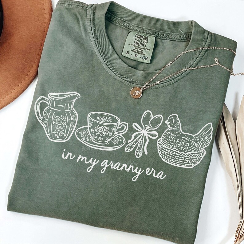 Puede incluir: Camiseta verde salvia con un gr&aacute;fico blanco de una jarra, una taza de t&eacute;, cucharas y una gallina en una cesta. El texto "in my granny era" est&aacute; debajo. Un collar dorado con un colgante est&aacute; sobre la camiseta.