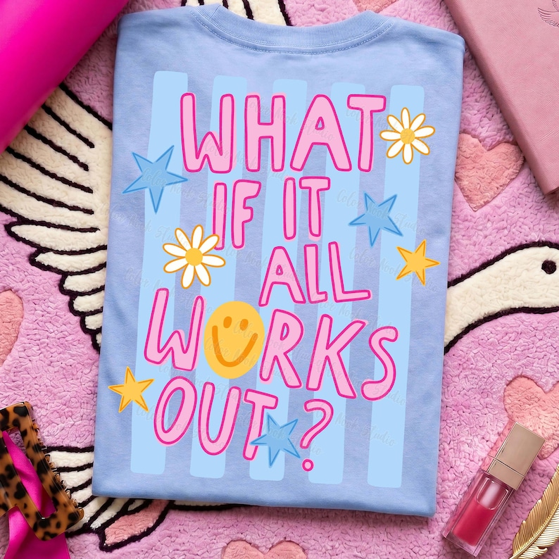 May include: Light blue t-shirt with the text "WHAT IF IT ALL WORKS OUT?" in pink, with a yellow smiley face, stars, and flowers. The shirt is on a pink and white patterned surface.