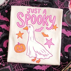 Może przedstawiać: Kremowa koszulka z napisem "JUST A SPOOKY" w kolorze r&oacute;żowym. Koszulka przedstawia kresk&oacute;wkową gęś, dynię, nietoperze i gwiazdy w odcieniach r&oacute;żu i pomarańczy, idealna na Halloween.