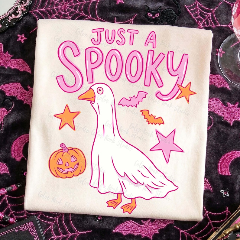 Może przedstawiać: Kremowa koszulka z napisem "JUST A SPOOKY" w kolorze r&oacute;żowym. Koszulka przedstawia kresk&oacute;wkową gęś, dynię, nietoperze i gwiazdy w odcieniach r&oacute;żu i pomarańczy, idealna na Halloween.