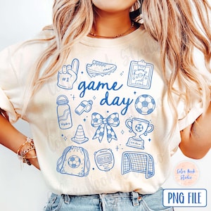 May include: A white t-shirt with a blue graphic design that says "game day" and features various sports-related icons, including a football, a whistle, a trophy, a football boot, and a water bottle.