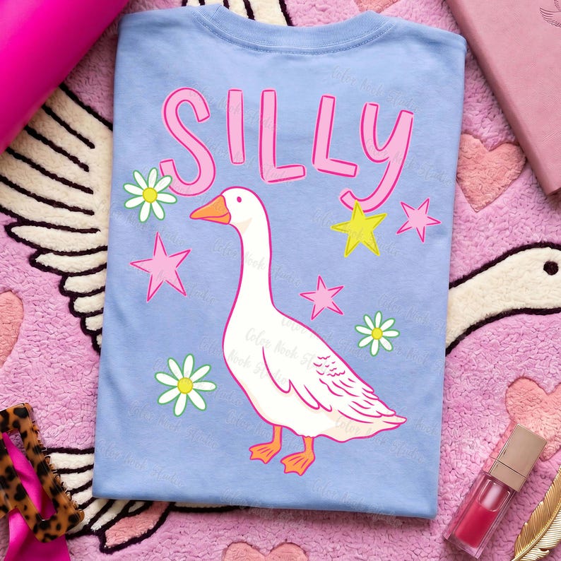 May include: Light blue t-shirt with the word "SILLY" in pink, a white goose illustration, pink stars, yellow stars, and white daisies. The shirt is a casual, fun design.