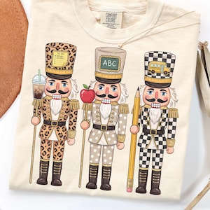 Könnte beinhalten: Ein cremefarbenes T-Shirt mit einer Grafik von drei Nussknackern. Der erste trägt eine Uniform mit Leopardenmuster und hält einen Eiskaffee. Der zweite hat eine gepunktete Uniform und hält einen Apfel. Der dritte hat eine karierte Uniform und hält einen Bleistift.
