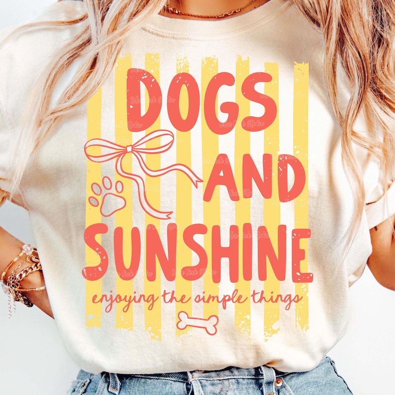 May include: Cream-colored t-shirt with a graphic design featuring the words "DOGS AND SUNSHINE" in coral letters. The design includes a paw print, a bow, and a bone, set against yellow and white vertical stripes. The phrase "enjoying the simple things" is also included.