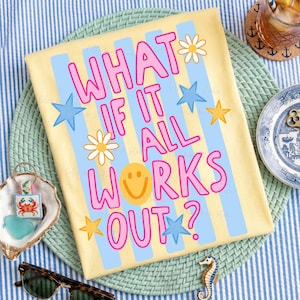 May include: Yellow t-shirt with the text "WHAT IF IT ALL WORKS OUT?" in pink, blue, and yellow. The design includes stars, daisies, and a smiley face. The shirt is on a blue and white striped surface.