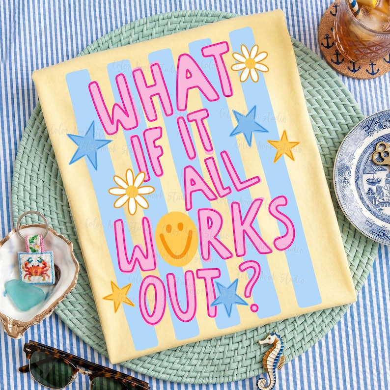May include: Yellow t-shirt with the text "WHAT IF IT ALL WORKS OUT?" in pink, blue, and yellow. The design includes stars, daisies, and a smiley face. The shirt is on a blue and white striped surface.