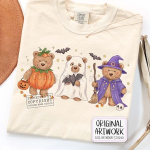 Op de afbeelding: Een cr&egrave;mekleurig t-shirt met een afbeelding van drie teddyberen in Halloween-kostuums: &eacute;&eacute;n als pompoen, &eacute;&eacute;n als spook met een vleermuis, en &eacute;&eacute;n als tovenaar met een bezem en hoed. Het ontwerp bevat kleine sterren, vleermuizen en snoepma&iuml;s.