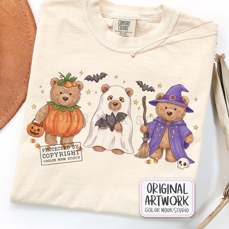 Op de afbeelding: Een cr&egrave;mekleurig t-shirt met een afbeelding van drie teddyberen in Halloween-kostuums: &eacute;&eacute;n als pompoen, &eacute;&eacute;n als spook met een vleermuis, en &eacute;&eacute;n als tovenaar met een bezem en hoed. Het ontwerp bevat kleine sterren, vleermuizen en snoepma&iuml;s.