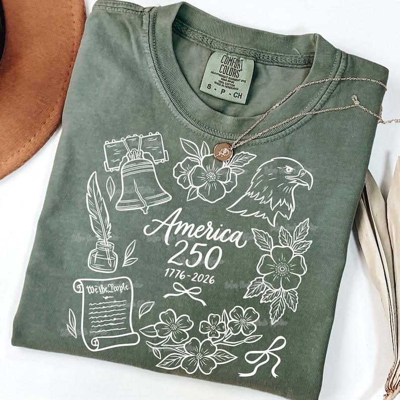 May include: Olive green t-shirt with a white graphic design featuring the Liberty Bell, an eagle, flowers, and the text "America 250 1776-2026". A gold necklace with a pendant is draped on the shirt.