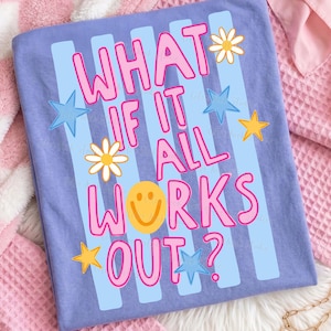 May include: A light purple t-shirt with the phrase "WHAT IF IT ALL WORKS OUT?" in pink lettering. The design includes blue stripes, yellow stars, white daisies, and a yellow smiley face.