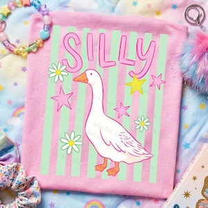 May include: Pink t-shirt with the word "SILLY" in pink letters above a white goose illustration. The shirt has green vertical stripes and is decorated with pink stars and white daisies.