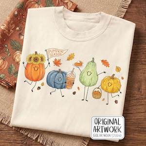May include: A cream-colored t-shirt featuring a graphic of four anthropomorphic pumpkins with smiling faces. One holds a flag that reads PUMPKIN SEASON, another holds a coffee cup and a slice of pie, and one carries a small basket of pumpkins.