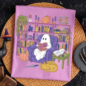 May include: A lavender graphic print showing a ghost reading in a purple armchair in front of a bookshelf. The bookshelf is filled with books, candles, and a carved pumpkin. A golden dragon and a knife are at the bottom.