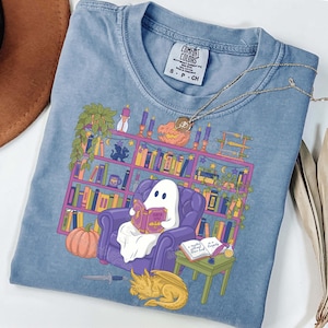 May include: Pale blue Comfort Colors t-shirt with a graphic of a ghost reading in a purple armchair in front of a bookshelf. A pumpkin, dragon, and table with a book are also shown. The shirt has the Comfort Colors label.
