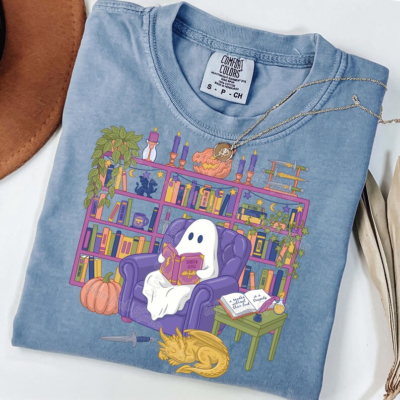 May include: Pale blue Comfort Colors t-shirt with a graphic of a ghost reading in a purple armchair in front of a bookshelf. A pumpkin, dragon, and table with a book are also shown. The shirt has the Comfort Colors label.