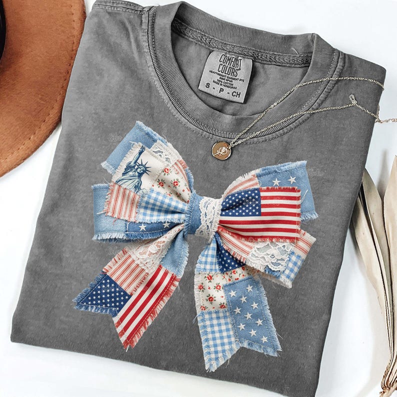 May include: Grey Comfort Colors t-shirt with a patriotic bow design. The bow features patchwork fabric with American flags, stars, and Liberty illustrations. A silver necklace with a pendant is also visible.