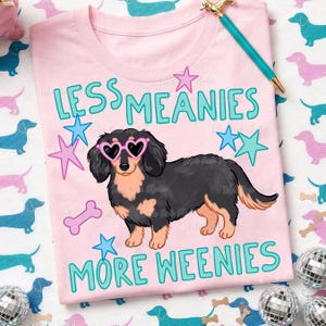 Op de afbeelding: Lichtroze T-shirt met een cartoon teckel met een hartvormige zonnebril. De tekst "LESS MEANIES MORE WEENIES" is turquoise. Roze en turquoise sterren en een botgrafiek omringen de hond.