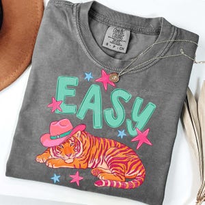 Puede incluir: Una camiseta gris Comfort Colors con la palabra "EASY" en letras turquesas y acentos de estrellas rosas. Un tigre con un sombrero de vaquero rosa est&aacute; debajo de la palabra. Un collar plateado con un peque&ntilde;o colgante est&aacute; en la camisa.