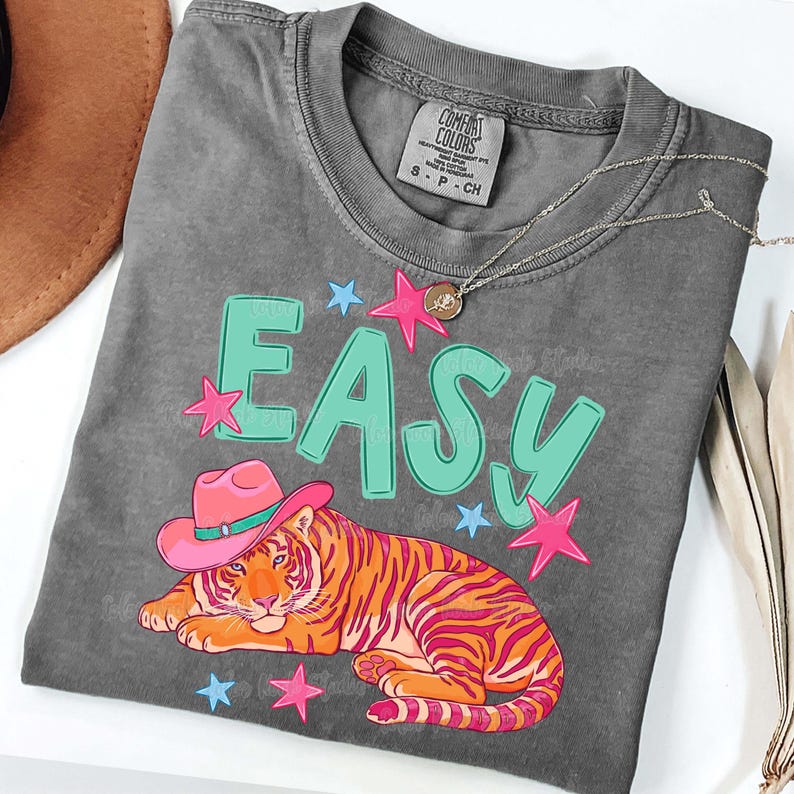 Puede incluir: Una camiseta gris Comfort Colors con la palabra "EASY" en letras turquesas y acentos de estrellas rosas. Un tigre con un sombrero de vaquero rosa est&aacute; debajo de la palabra. Un collar plateado con un peque&ntilde;o colgante est&aacute; en la camisa.