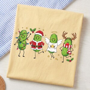 May include: A light yellow t-shirt featuring a graphic of four cartoon pickles. One is wrapped in colourful Christmas lights, one wears a Santa suit, one is dressed as an angel with wings and a halo, and one wears reindeer antlers and a red nose.