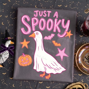 Może przedstawiać: Szara tkanina z napisem "JUST A SPOOKY" w kolorze r&oacute;żowym. Przedstawiono białego gęsia w przebraniu ducha, a także pomarańczowe gwiazdy, nietoperze i dynię. Projekt ma motyw Halloween.