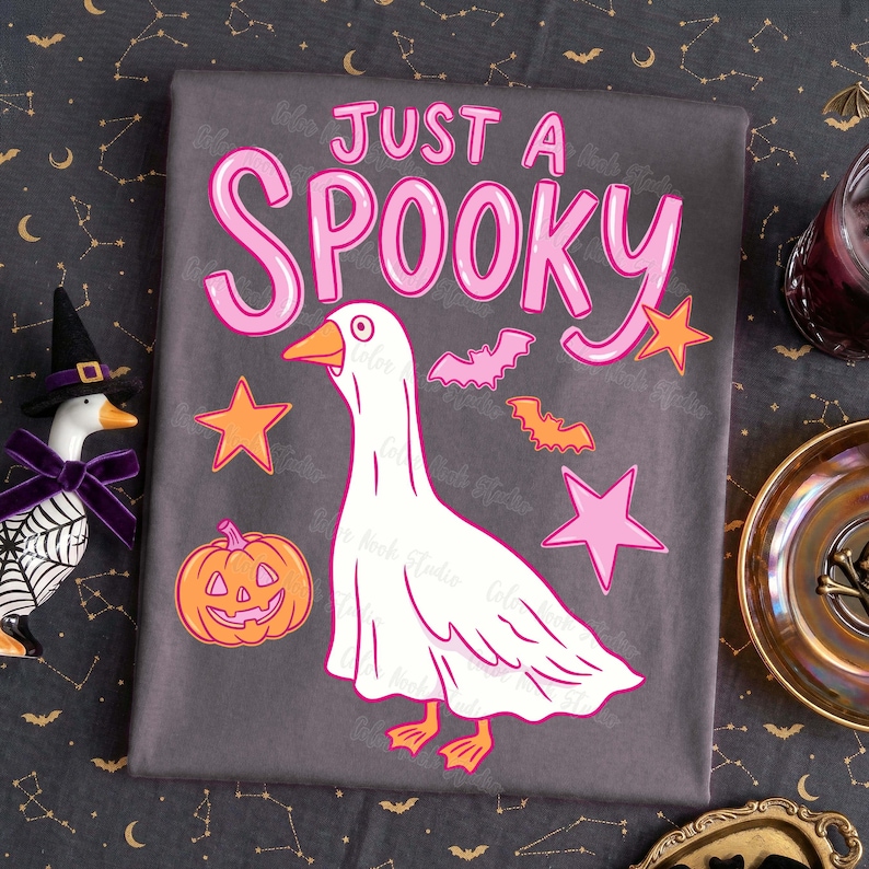 Może przedstawiać: Szara tkanina z napisem "JUST A SPOOKY" w kolorze r&oacute;żowym. Przedstawiono białego gęsia w przebraniu ducha, a także pomarańczowe gwiazdy, nietoperze i dynię. Projekt ma motyw Halloween.