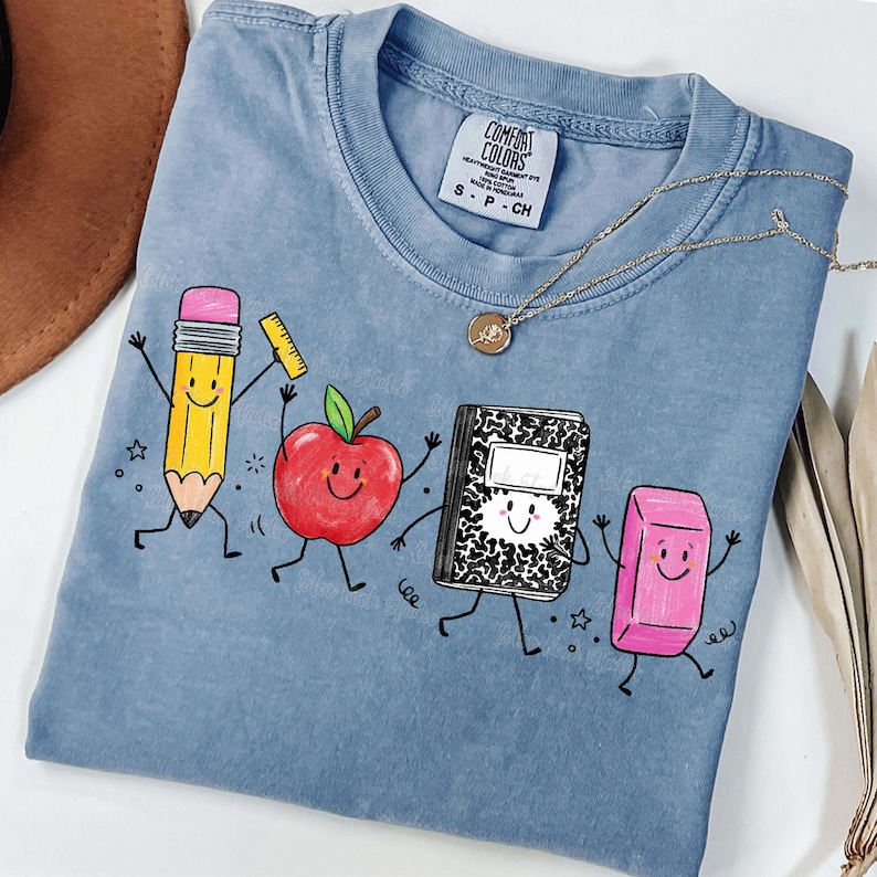 May include: A light blue Comfort Colors t-shirt featuring a playful graphic of animated school supplies: a pencil, apple, notebook, and eraser. The shirt has a crew neck and short sleeves. A gold necklace is draped across the shirt.